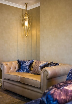 A beige leather sofa with patterned cushions, a hanging gold-colored light fixture, and a textured wall in a room with a checkered floor. - Arden House (Stratford-upon-Avon)