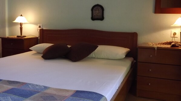 Una cama bien hecha con colcha escocesa, dos almohadas y un cabecero de madera. Hay dos mesitas de noche con lámparas, un pequeño reloj de pared y un aparador de madera con cajones.
