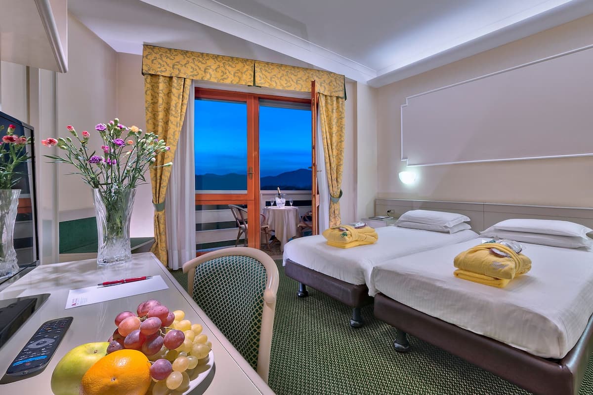 Een hotelkamer met twee bedden, een bureau met een computer en een bloevas, een eettafel met fruit, en uitzicht op de bergen door het raam.