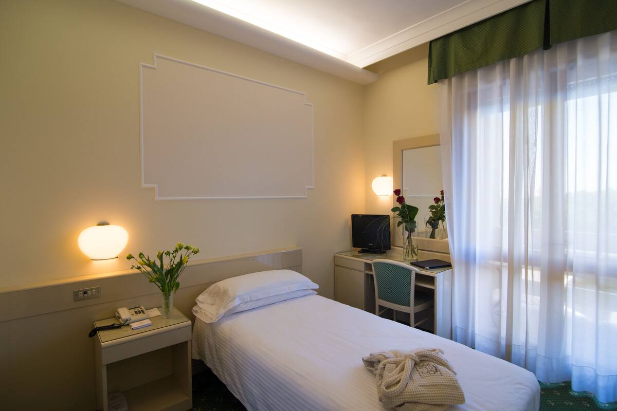 Een hotelkamer met een bed, een bureau met een computer, een stoel, een nachtkastje met een lamp en een raam met dunne gordijnen.