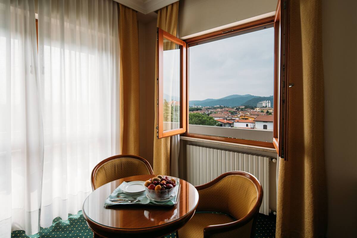 Hotelkamer met een eettafel voor twee personen, een fruitschaal en uitzicht op een stad en bergen door een raam.