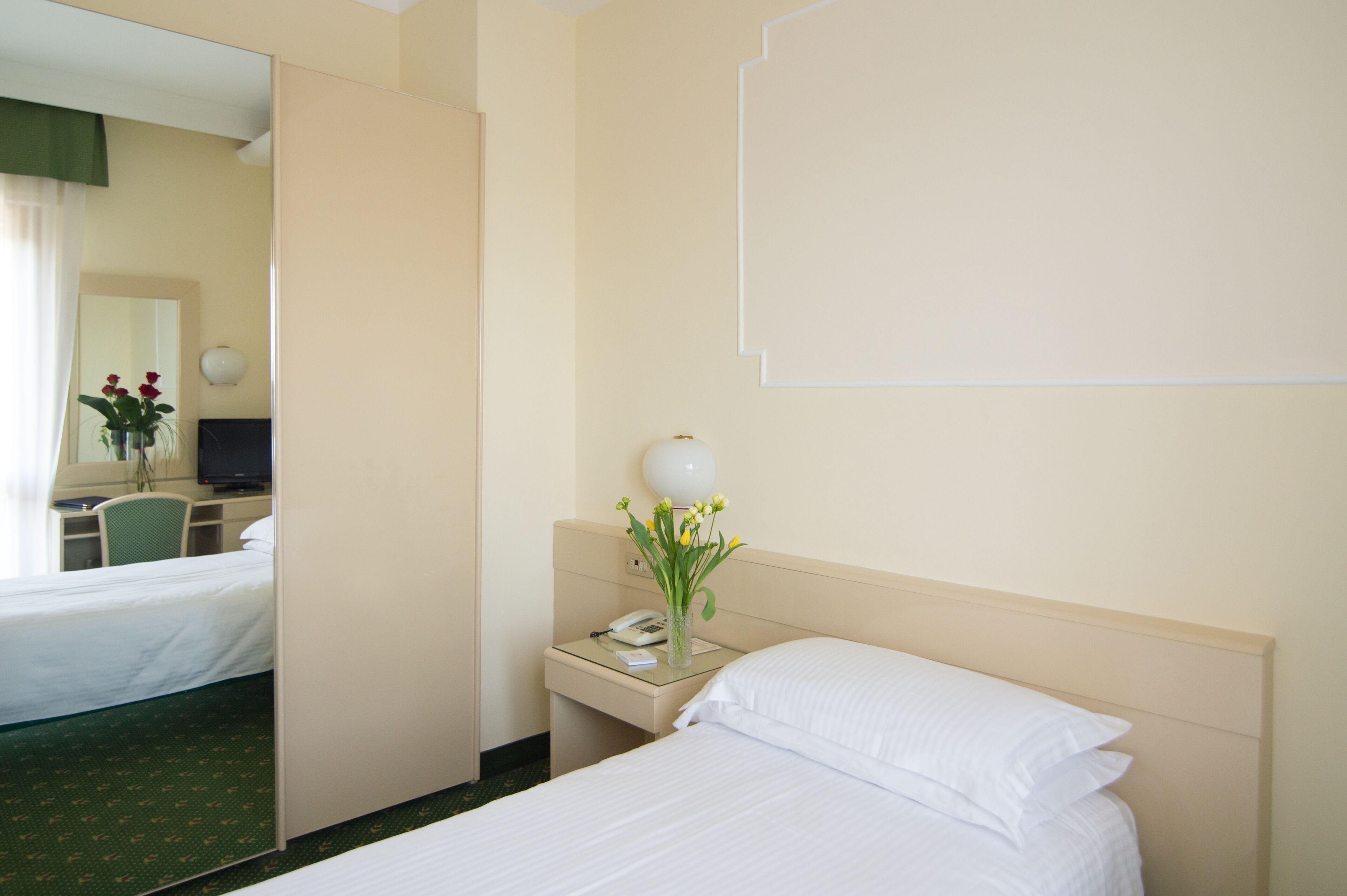 A hotel room with a bed, a bedside table with a lamp and a vase of flowers, a desk with a computer, and a mirror.