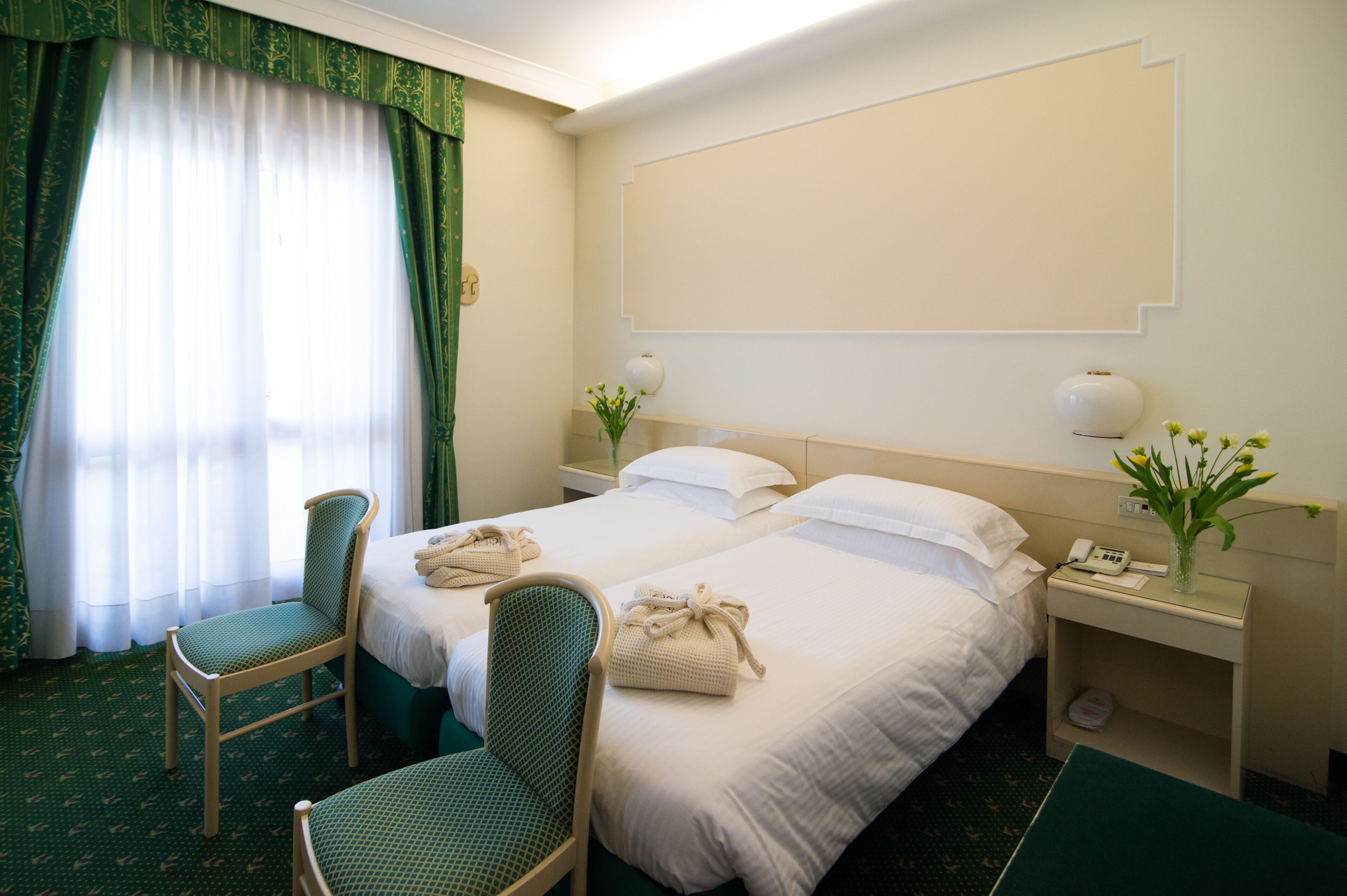 Habitación de hotel con dos camas, una silla, una mesita de noche con teléfono y florero, y una ventana con cortinas.