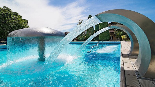 Piscina com um moderno detalhe aquático, rodeada por um deck revestido e vegetação.