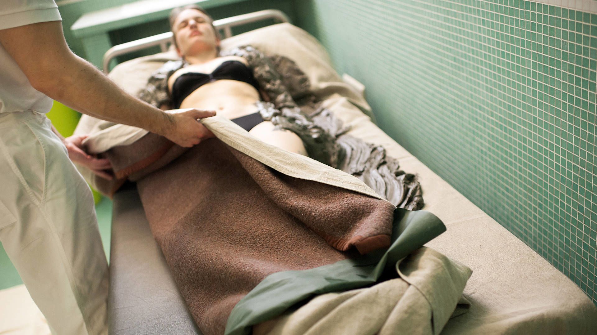 A hospital room with a patient lying on a bed, covered with a blanket, and a medical professional attending to them.
