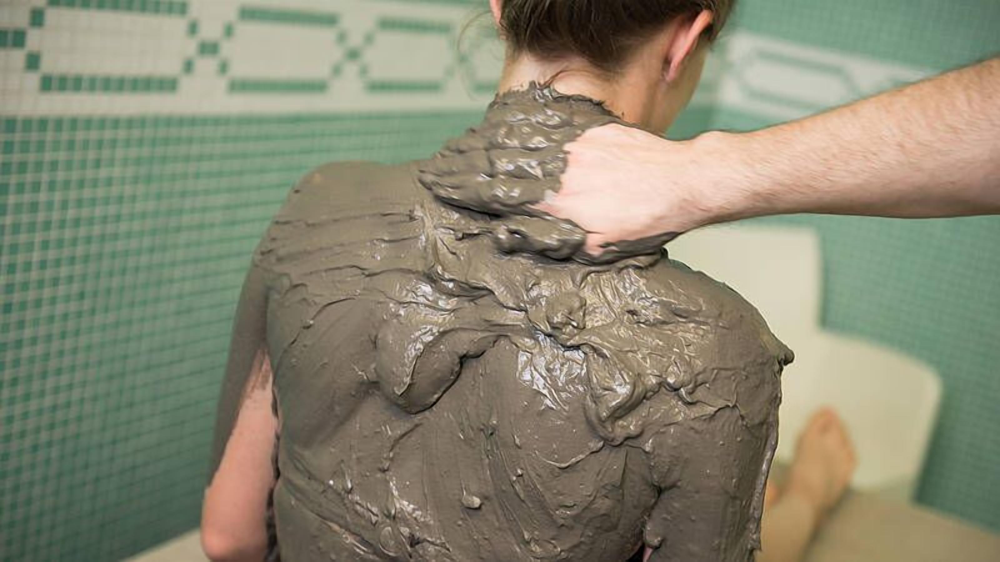 Una persona cubierta con una sustancia espesa y marrón, posiblemente barro o arcilla, en un baño con pared de azulejos verdes.