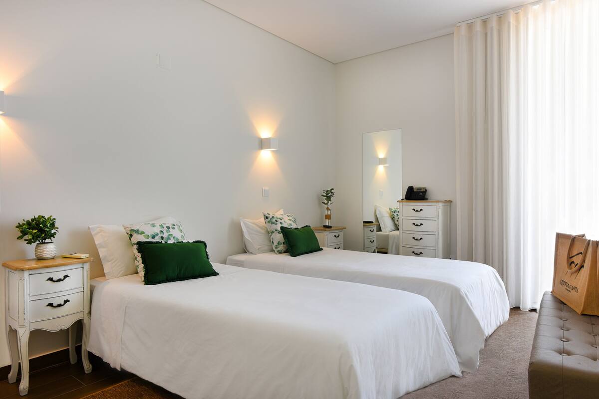 A hotel room with two beds, white bedding, a small bedside table with a plant, a mirror, a dresser, and a large window with curtains.