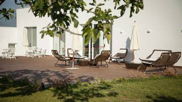 Een terras met stoelen en parasols, een wit gebouw op de achtergrond en een boom in de voorgrond.