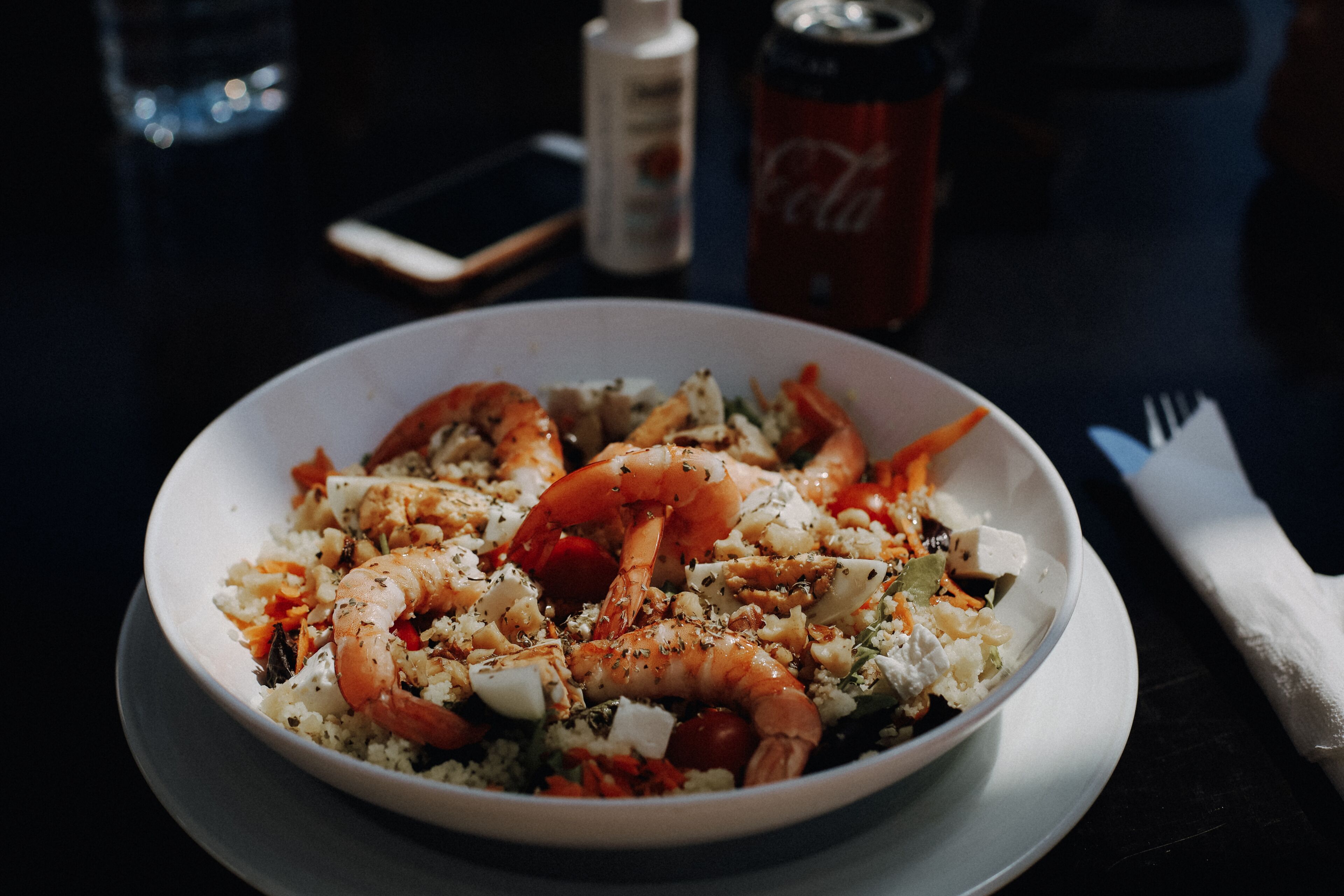 A bowl of shrimp and rice dish on a table with a bottle and a can in the background.