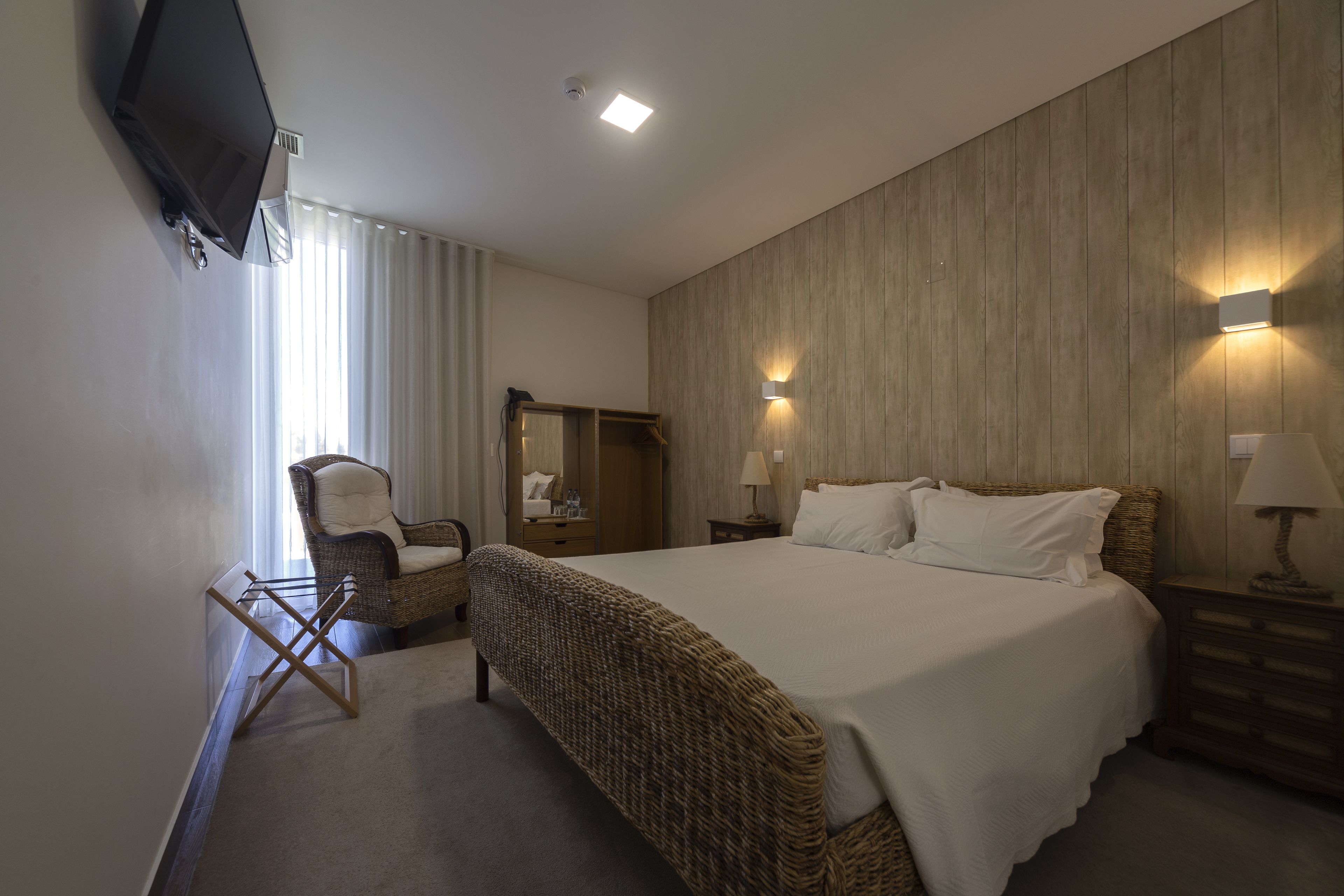 Een slaapkamer met een groot bed, een flatscreen televisie aan de muur, een rieten fauteuil, een houten garderobe en twee bedlampjes.