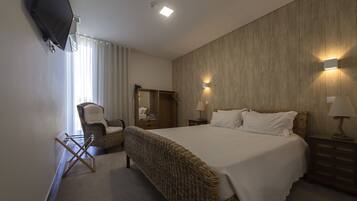 Een slaapkamer met een groot bed, een flatscreen televisie aan de muur, een rieten fauteuil, een houten garderobe en twee bedlampjes.