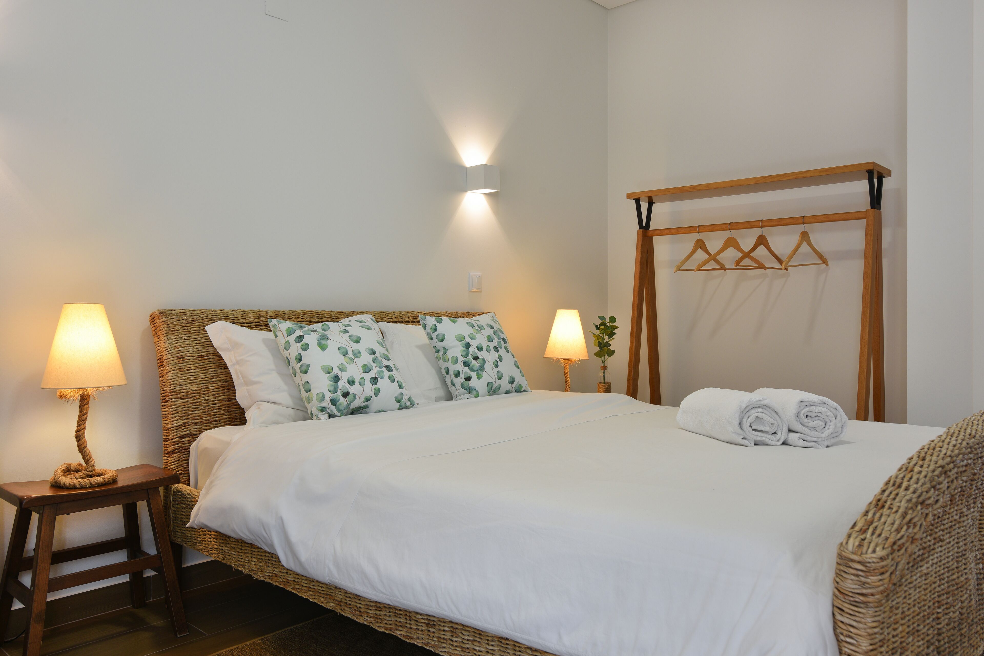 Una cama bien hecha con sábanas blancas y almohadas con estampado de hojas verdes, una lámpara de noche y una mesita de noche de madera.