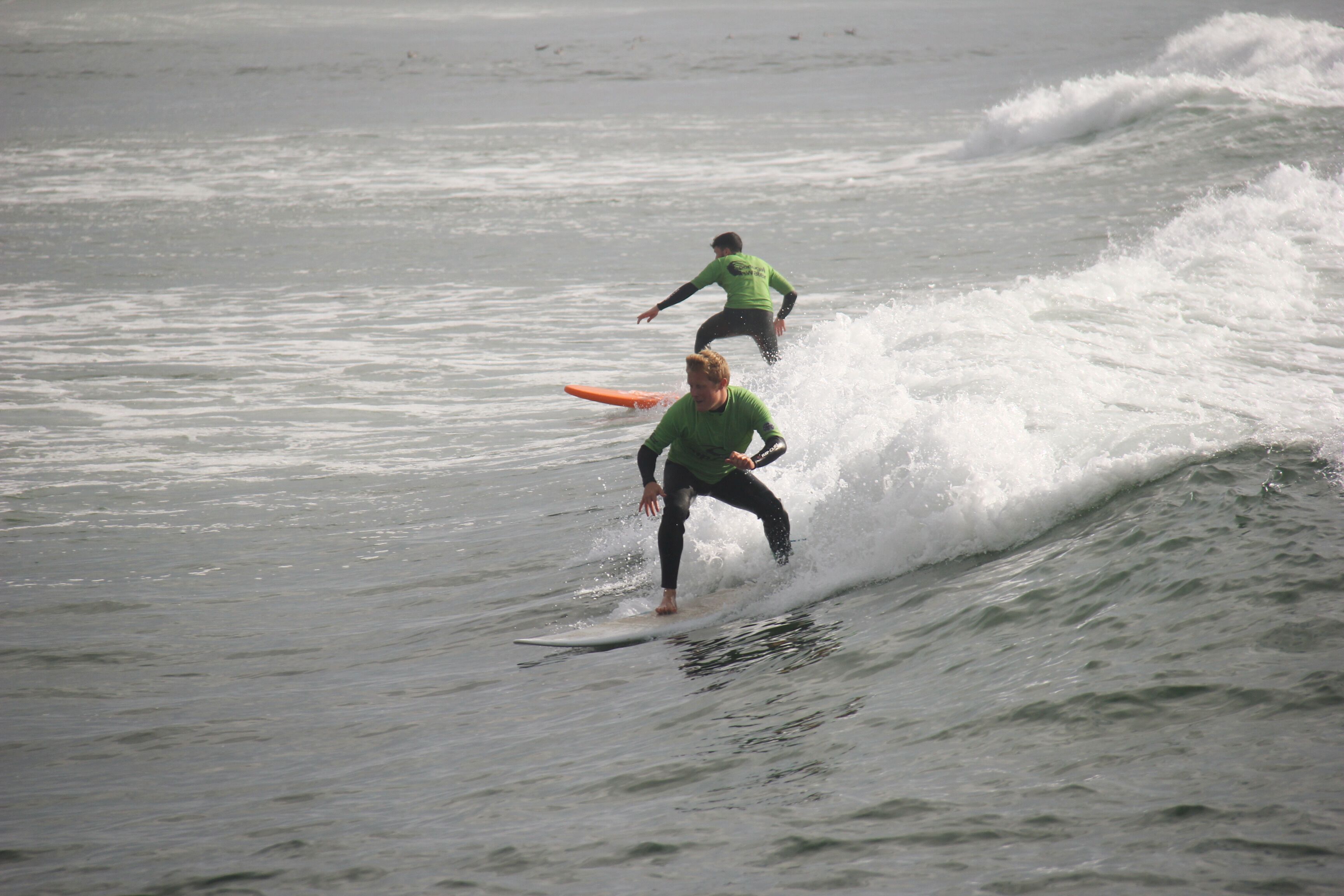 Deux surfeurs sur une vague, l’un portant un t-shirt vert et l’autre un t-shirt noir.