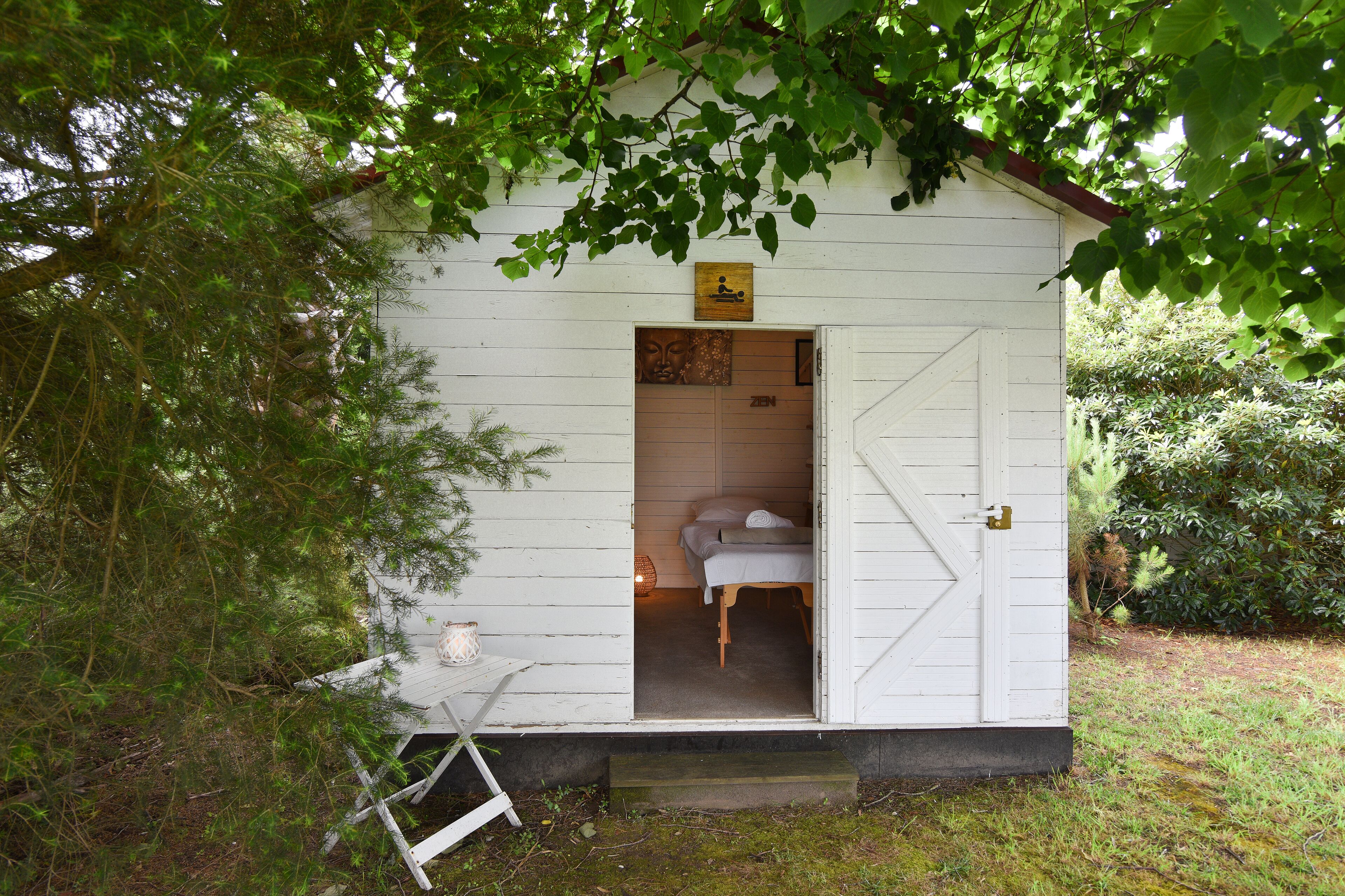Una cabaña de madera blanca y pequeña con una cama en su interior, una mesita con una vela, y un letrero sobre la puerta.