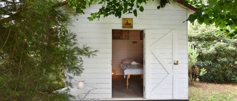 Una cabaña de madera blanca y pequeña con una cama en su interior, una mesita con una vela, y un letrero sobre la puerta.