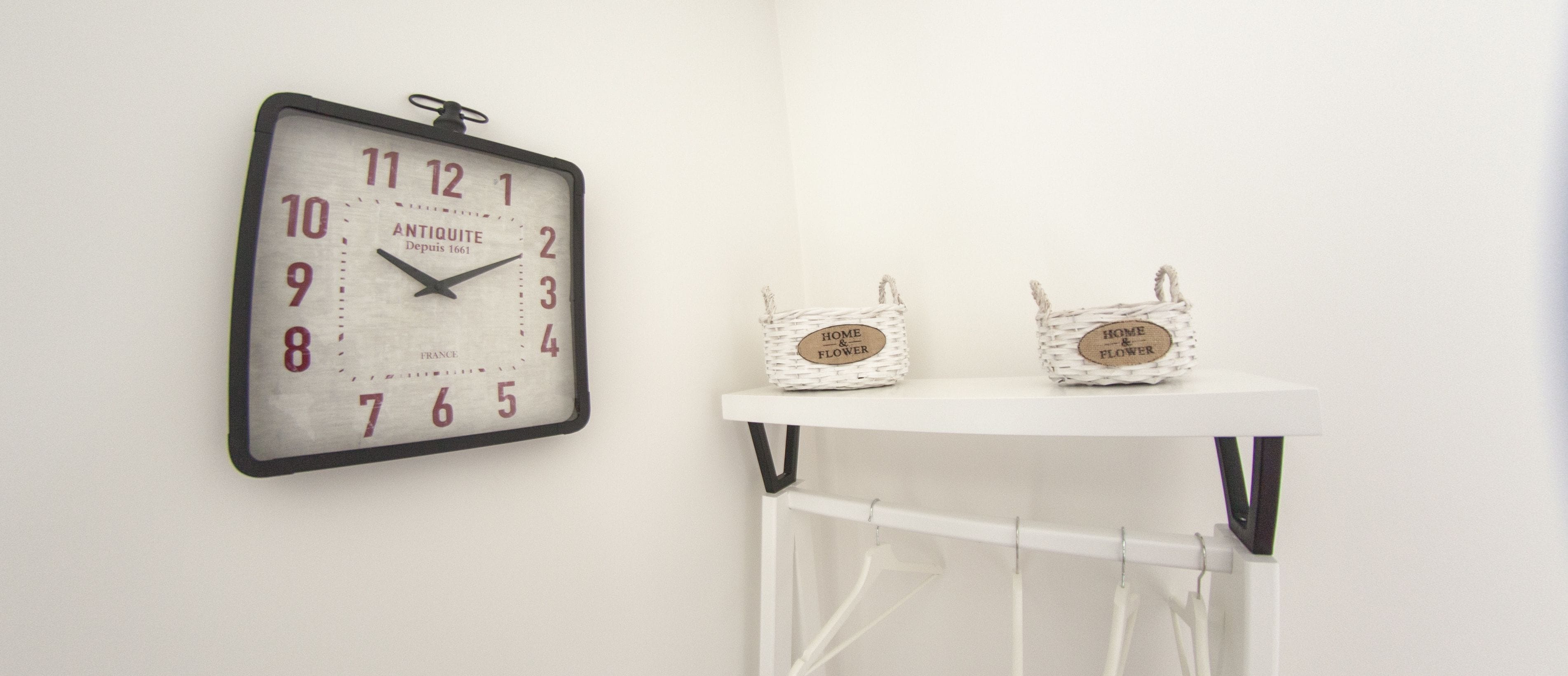 Een antieke klok aan de muur, met een witte wijzerplaat en zwarte cijfers, twee rieten manden met de tekst 'HOME & FLOWER', en een witte plank met hangende spullen.