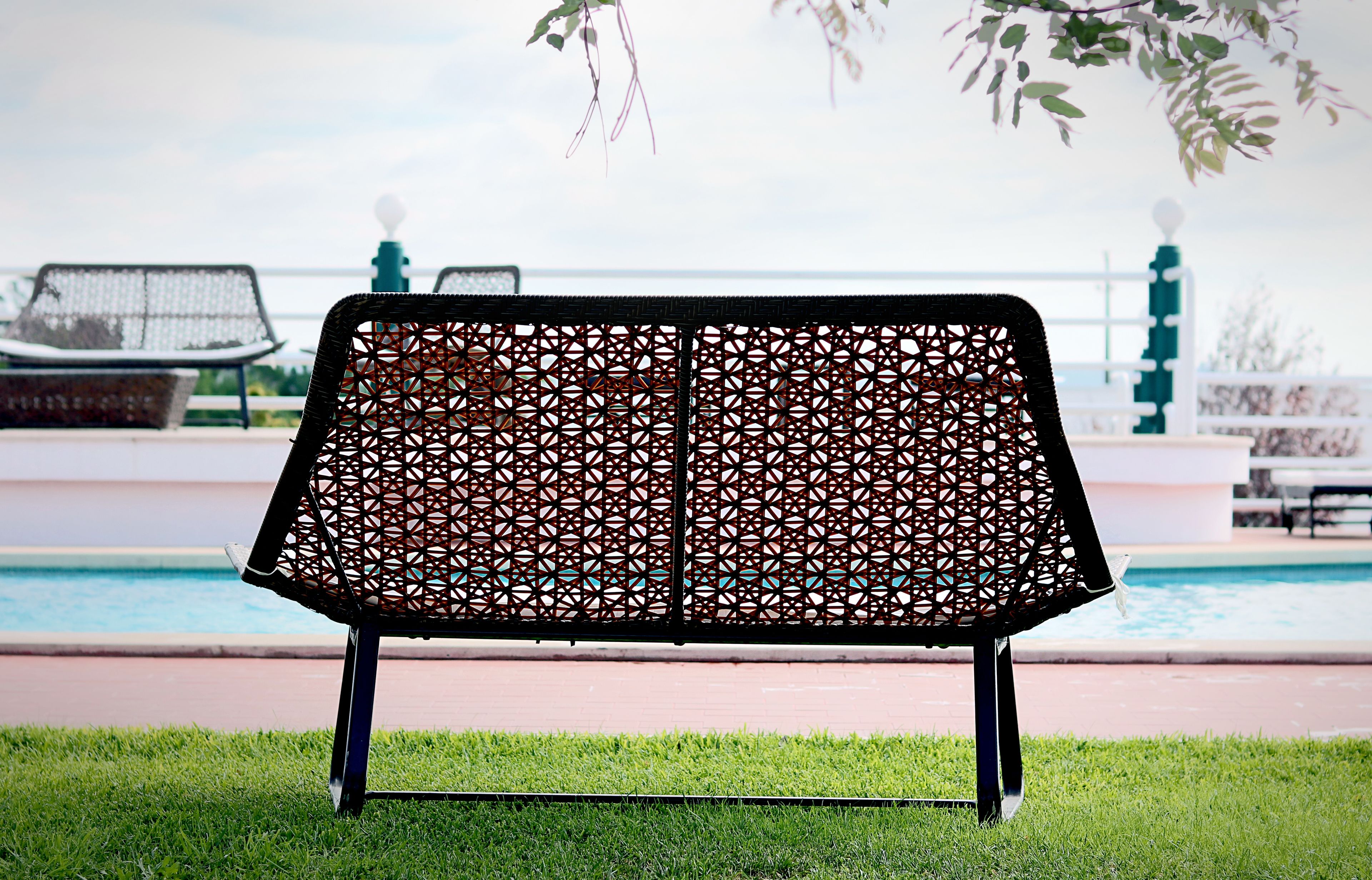 A bench with a geometric lattice design, positioned on grass near a poolside area.