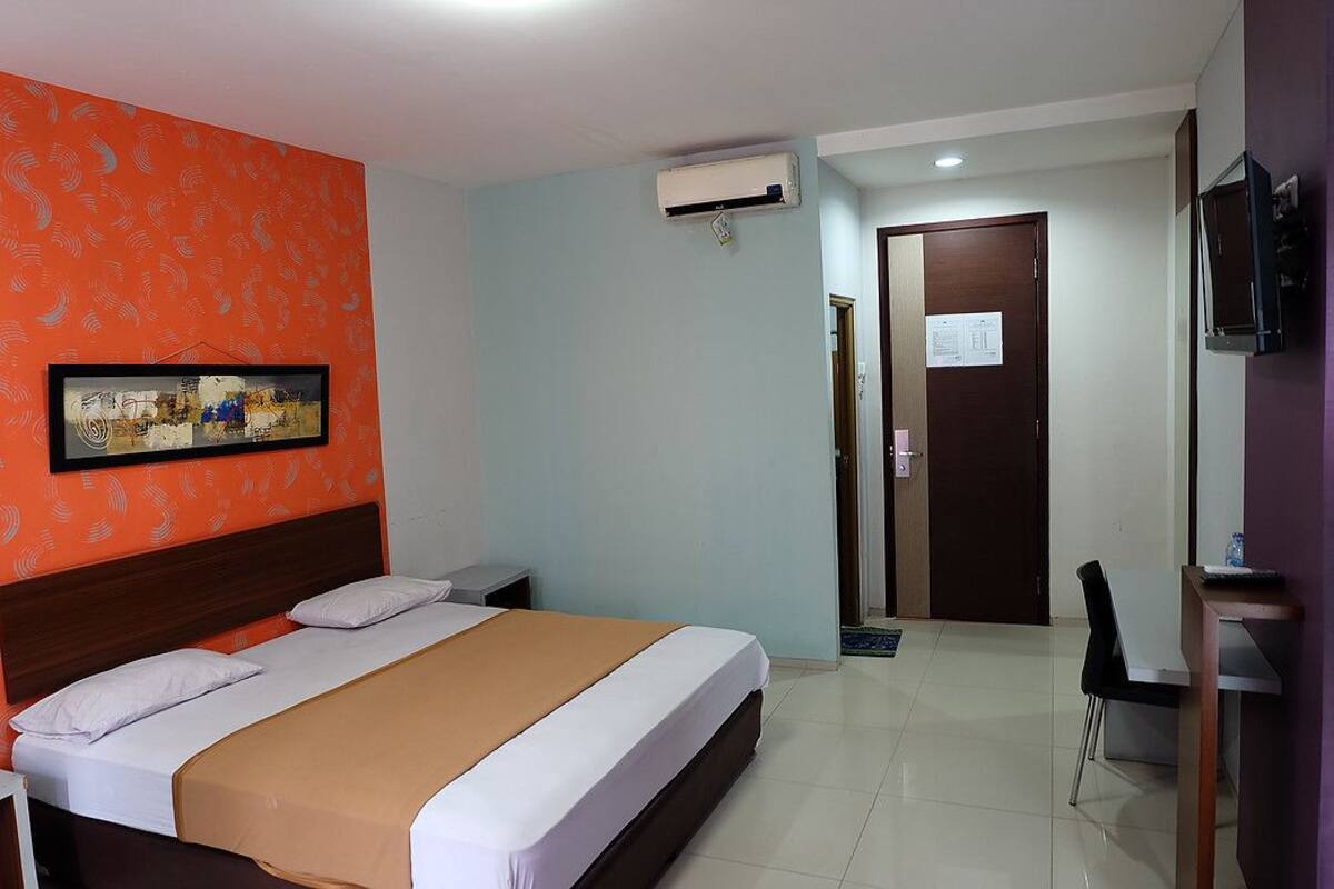 A hotel room with a bed, a desk, a chair, an air conditioning unit, and a television.