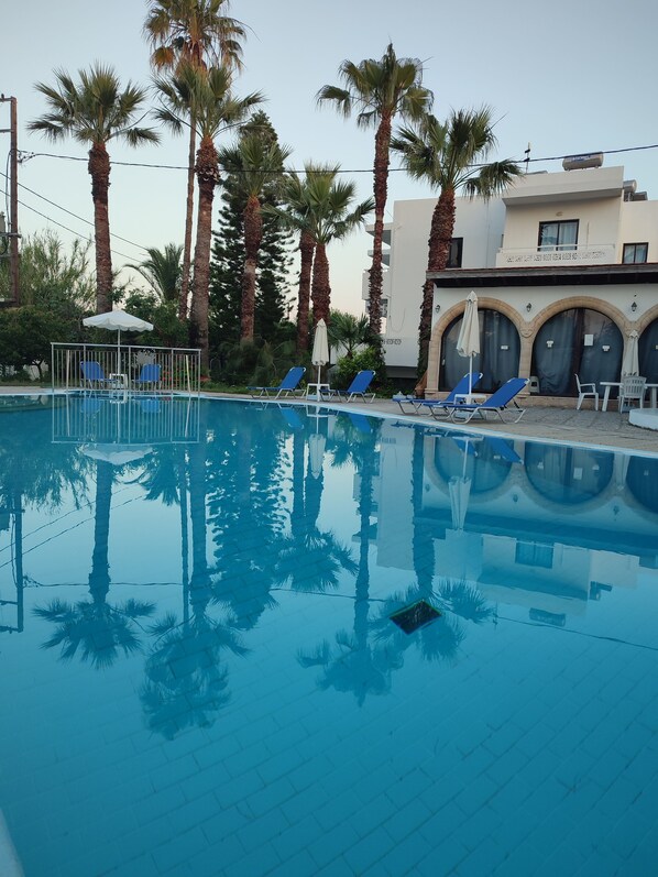 A swimming pool with blue water, surrounded by palm trees and lounge chairs. - Dolphin Apartments (Rhodes)