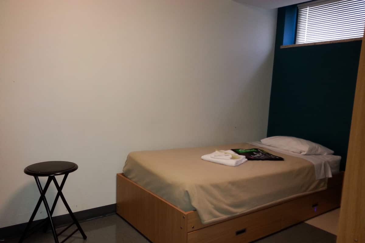 Ein kleines Zimmer mit einem Einzelbett, einem hölzernen Bettgestell, einem kleinen schwarzen Hocker und einem Fenster mit Jalousien.