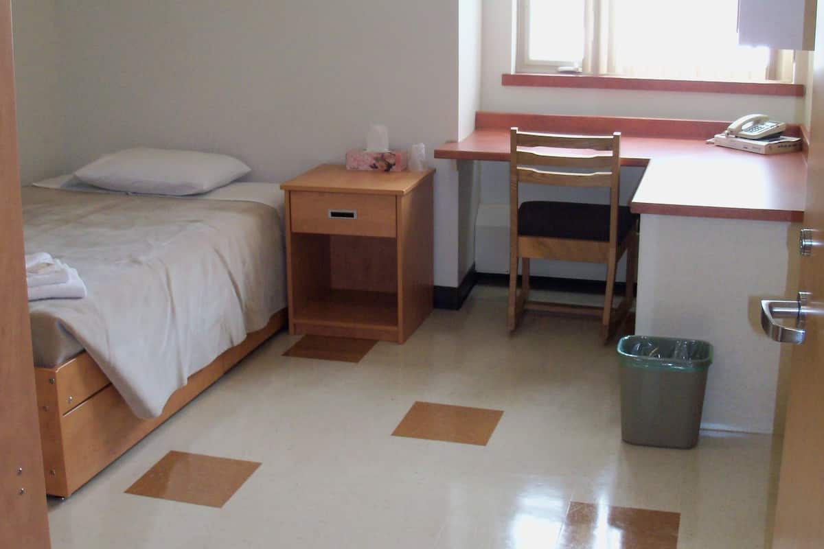 Ein kleines, sauberes Zimmer mit einem Bett, einem Schreibtisch und einem Stuhl. Es gibt ein Fenster mit Jalousien, einen Mülleimer und eine Tür.