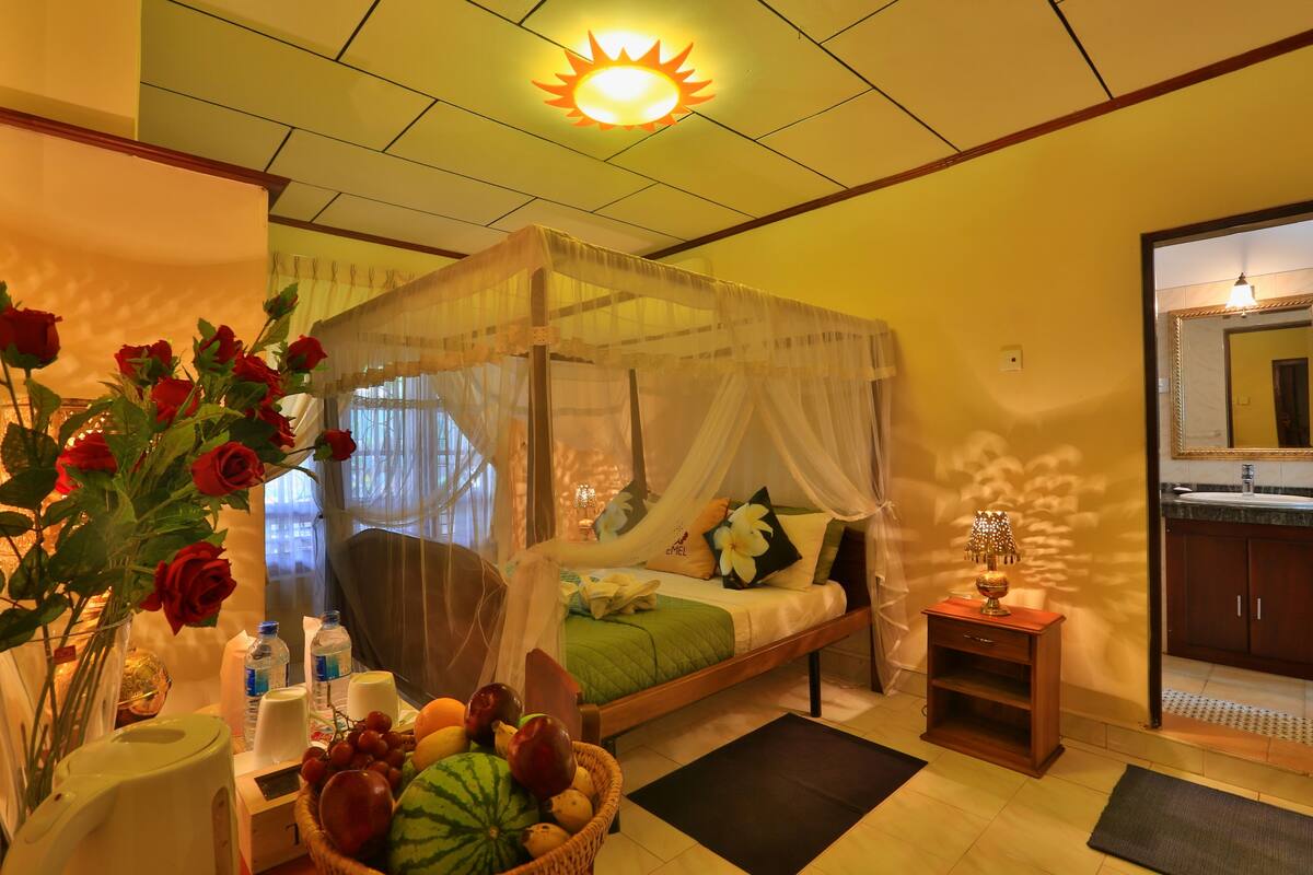 A room with a bed, a bedside table, a fruit basket, and a bathroom visible through an open door.