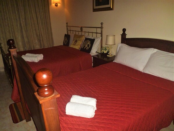 A hotel room with two beds, each with a wooden headboard, red bedding, and white pillows. There is a nightstand with a lamp and a vase of flowers. - Emel Boutique Hotel (Kochchikade)