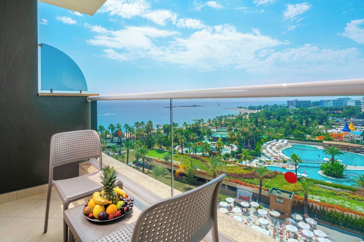 A balcony with a table and chairs, a fruit bowl, and a view of a resort with pools and greenery.