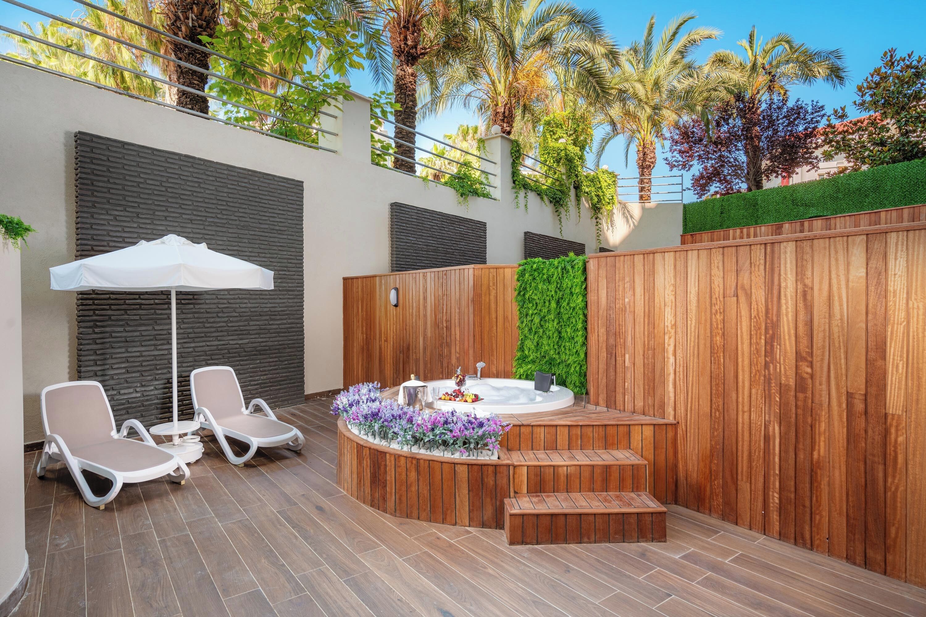 A modern outdoor patio with two lounge chairs, a white umbrella, a small table, and a hot tub.