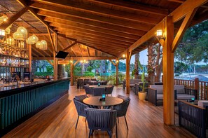 Un espace bar extérieur abrité, doté de poutres en bois, de tables rondes et de chaises, le tout entouré de verdure.