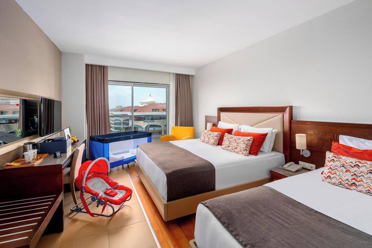 A hotel room with two beds, a desk with a computer, a TV, a window with curtains, and a view of buildings.