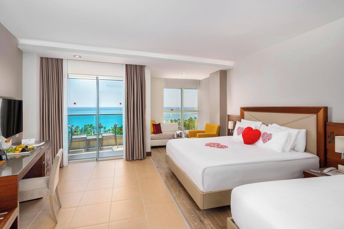 A hotel room with a balcony, a bed with white bedding, a TV, a desk with a chair, and a dining area with a table and chairs.