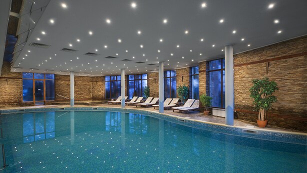 Una piscina interior con suelo de baldosas, varias tumbonas y grandes ventanales.