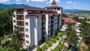 Un hotel de varios pisos con balcones, una estructura tipo torre y vistas a un pueblo y montañas al fondo.