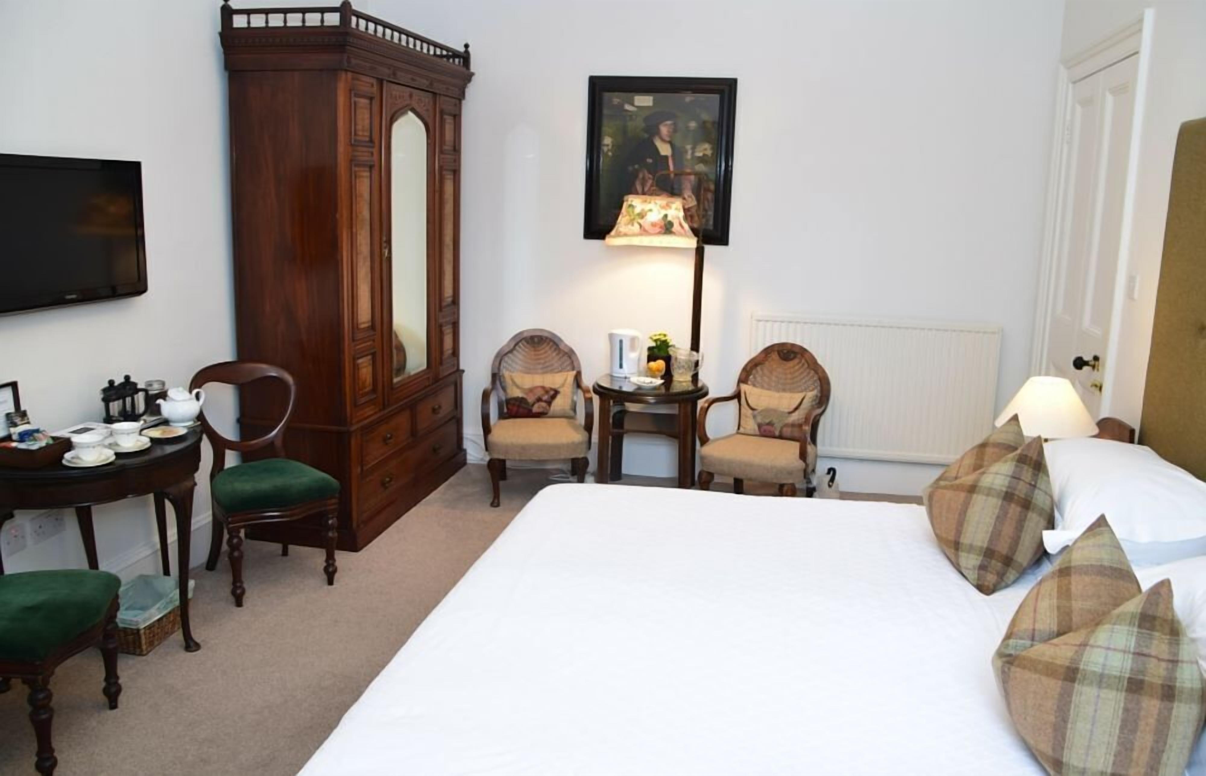 A hotel room with a large bed, two armchairs, a wooden cabinet, a small table with a lamp, and a painting on the wall.
