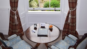 Una habitación con un ventanal grande, dos sillas, una mesita con un libro, un reloj y un frutero.