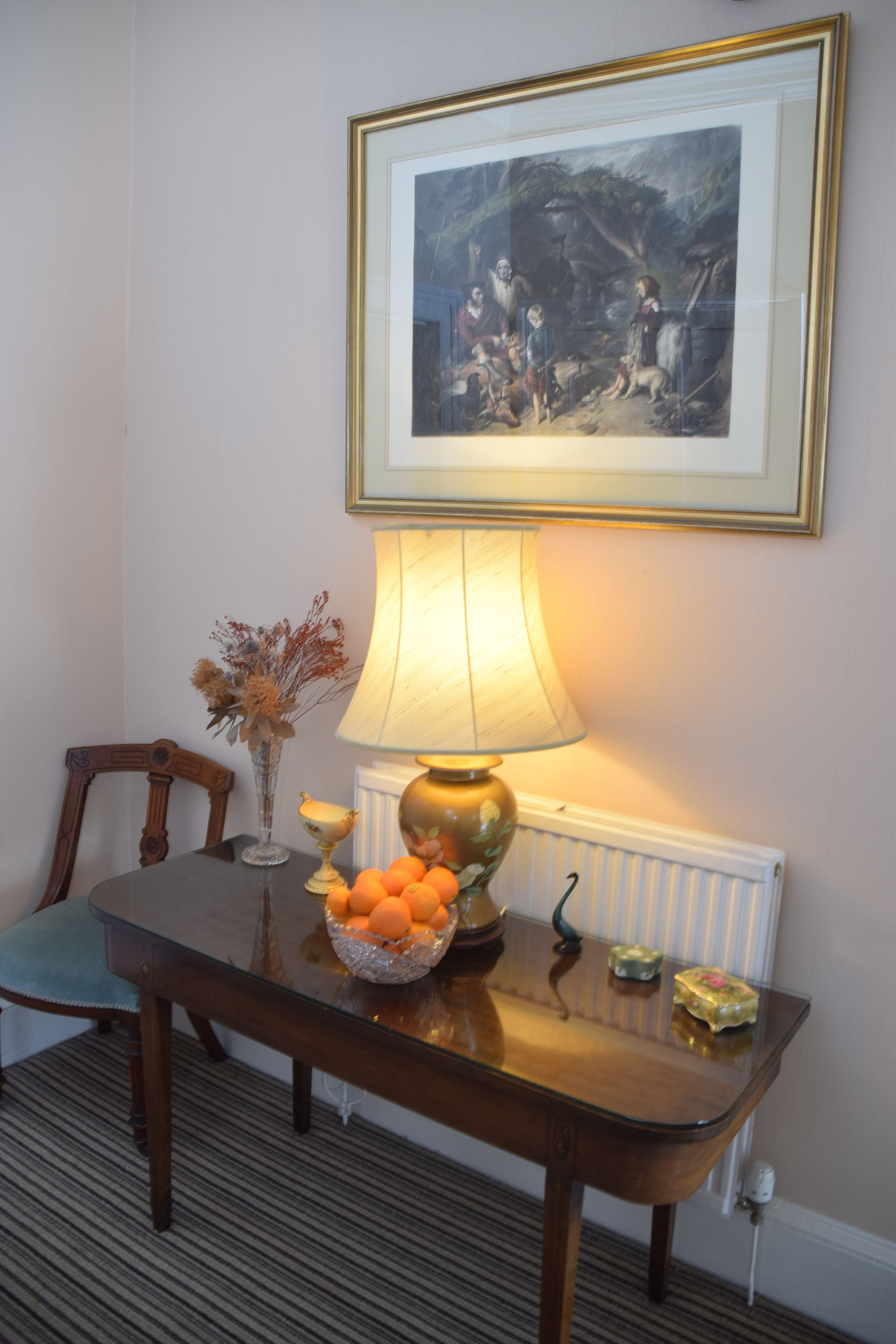 A dining table with a lamp, a framed picture, and a bowl of oranges.