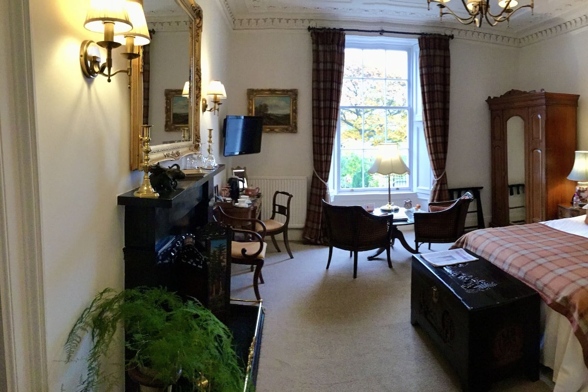 Habitación de hotel con cama, mesitas de noche, escritorio, silla, espejo y ventana con cortinas.