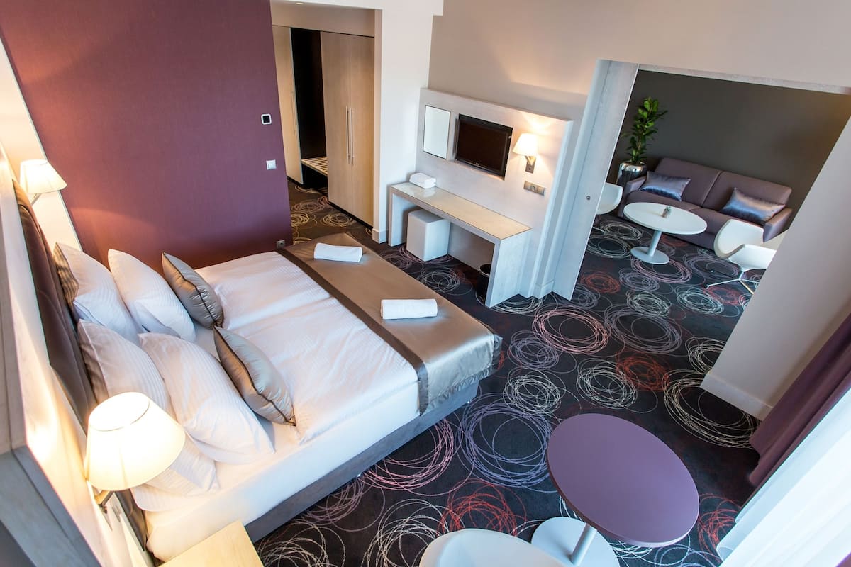 Habitación de hotel con cama, mesita de noche, lámpara, televisión, zona de estar con sofá y sillones, y una pequeña mesa.