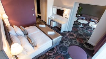 Habitación de hotel con cama, mesita de noche, lámpara, televisión, zona de estar con sofá y sillones, y una pequeña mesa.