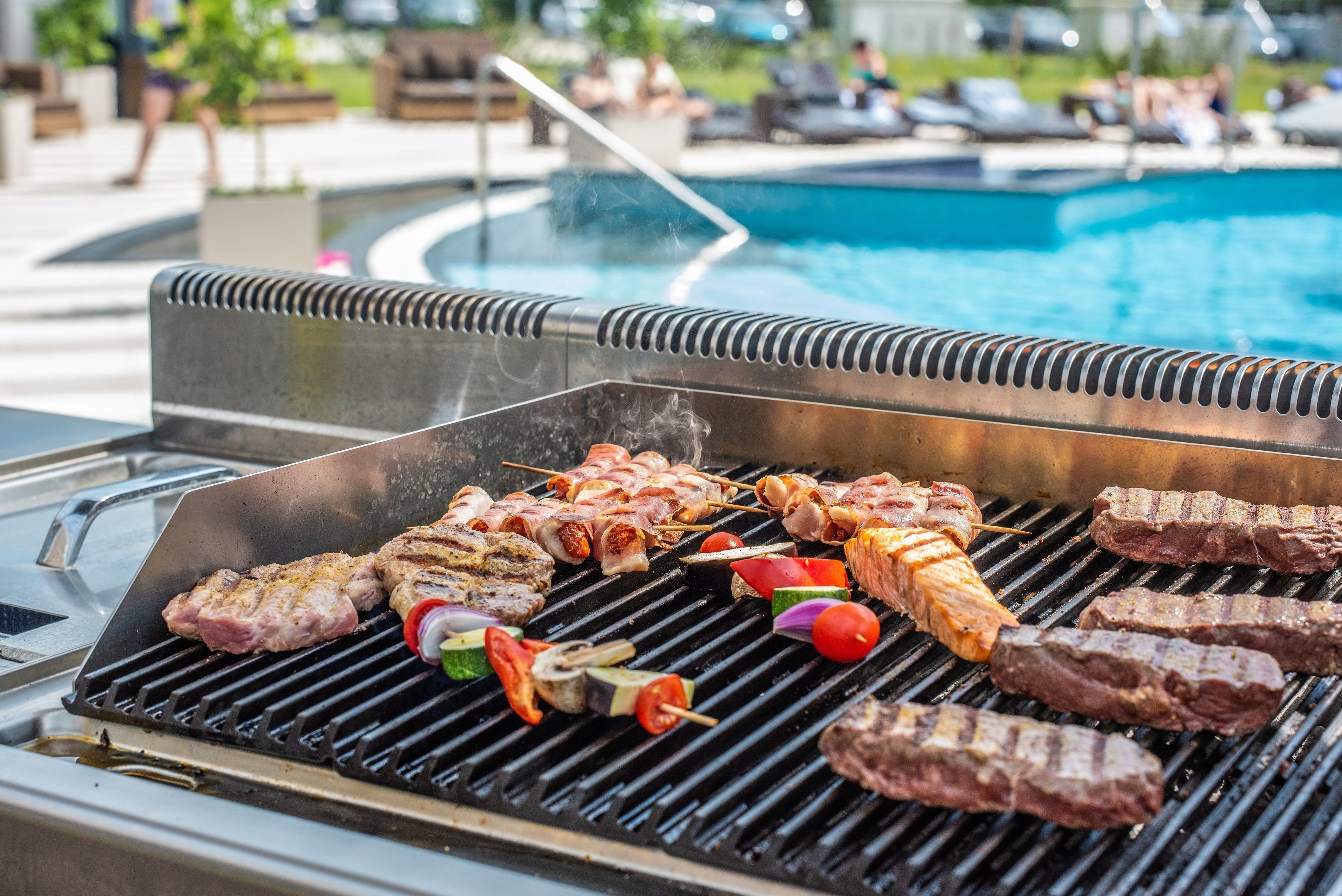 Una parrilla con diversos tipos de carne y verduras cocinándose al aire libre, cerca de la zona de la piscina.