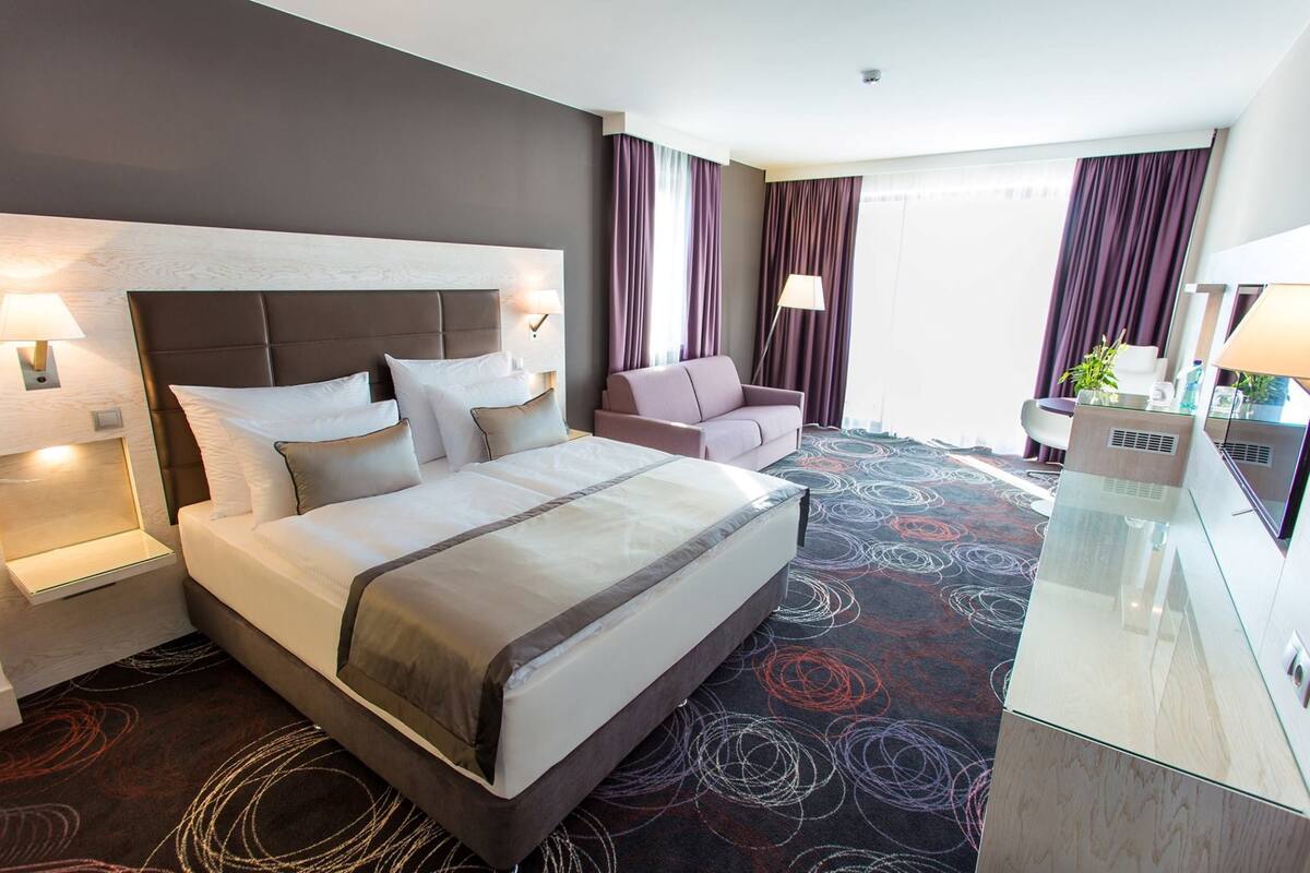 Habitación de hotel con una cama grande, un sofá, un escritorio con lámpara y una ventana con cortinas.
