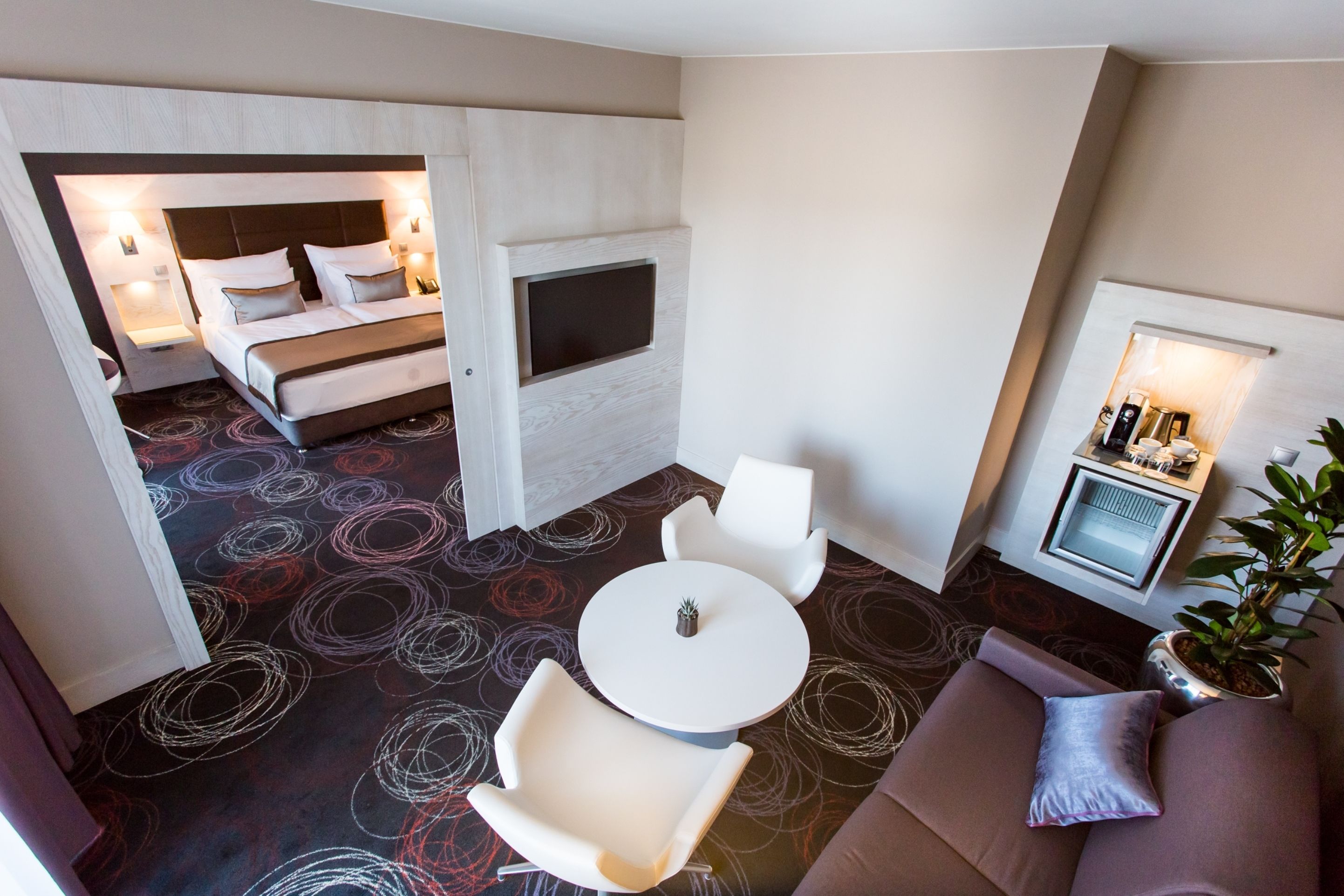 Quarto de hotel com cama, televisão, área de estar com mesa e cadeiras, e uma kitchenette.