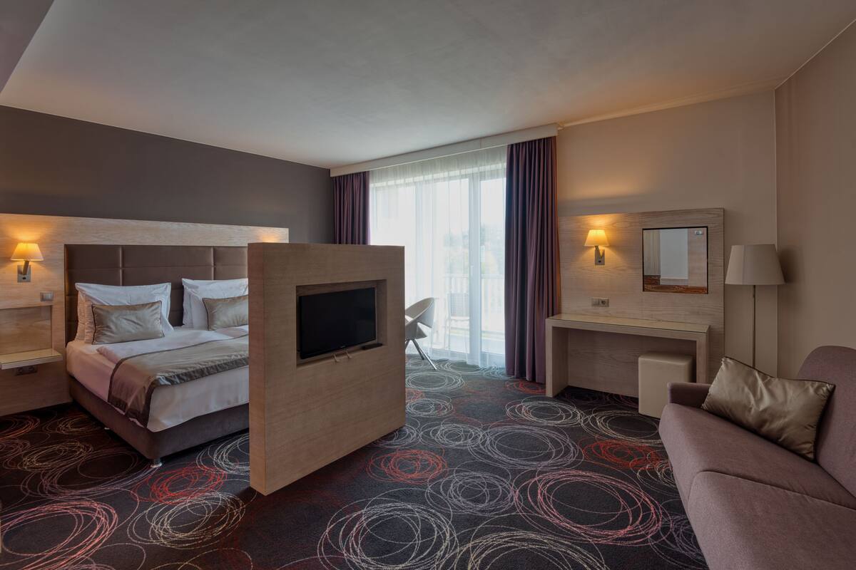 Una habitación de hotel con cama, escritorio, televisor, sofá y ventana con cortinas.