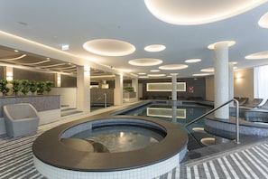 A modern indoor pool area with a hot tub, lounge chairs, and a tiled floor.