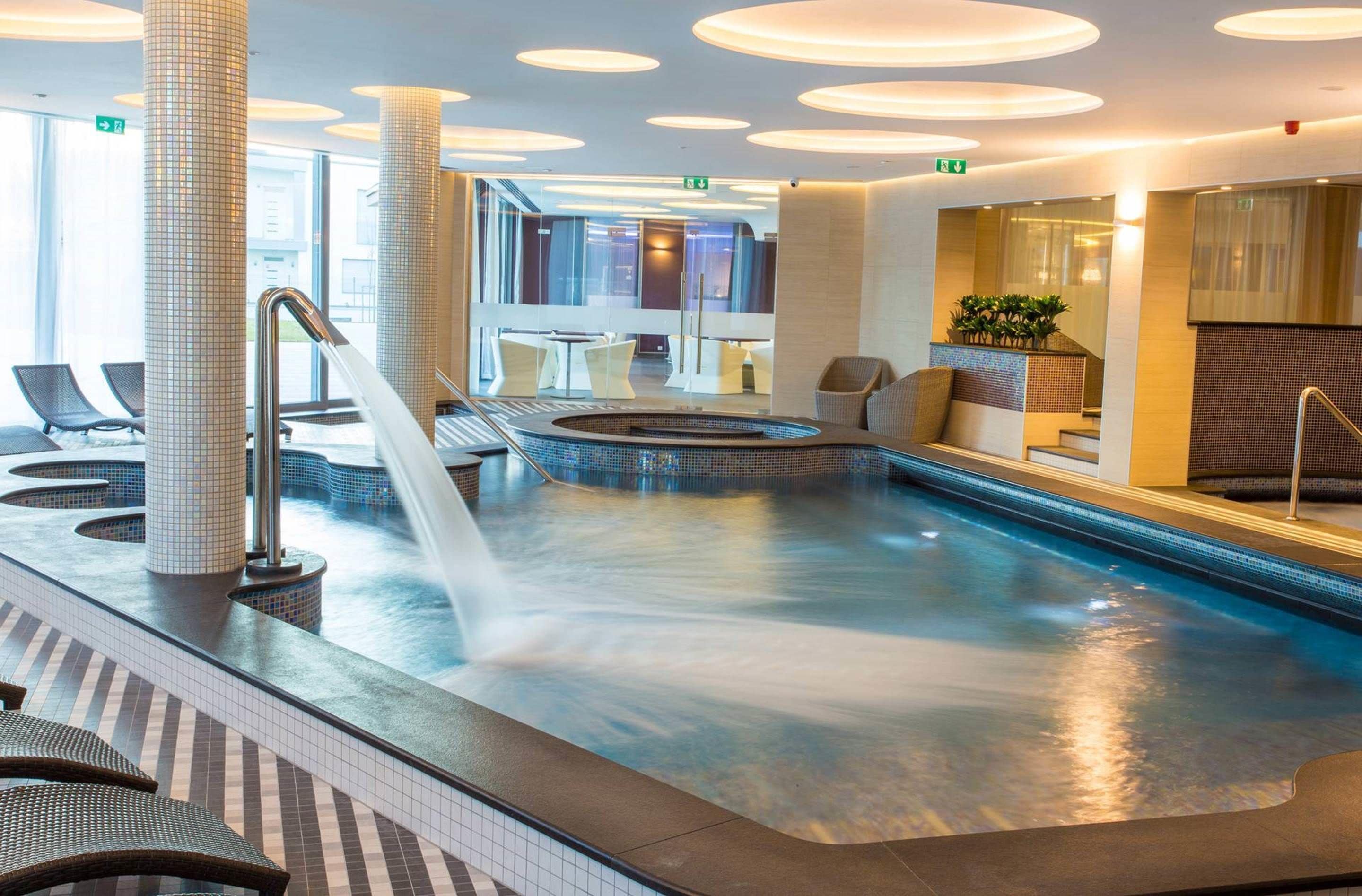 A modern indoor pool area with a central hot tub, seating areas, and a view of the adjacent restaurant.