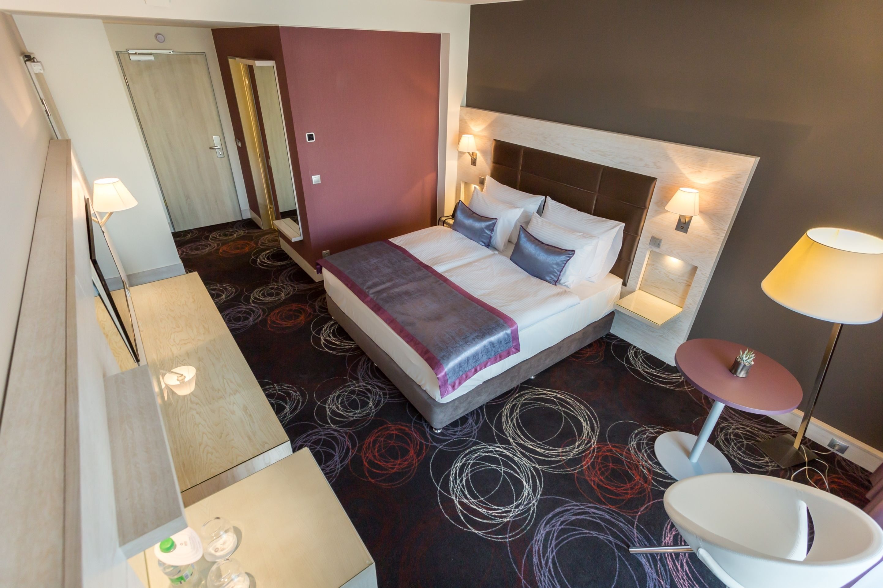 Quarto de hotel com cama, abajures, uma mesa redonda e um espelho.
