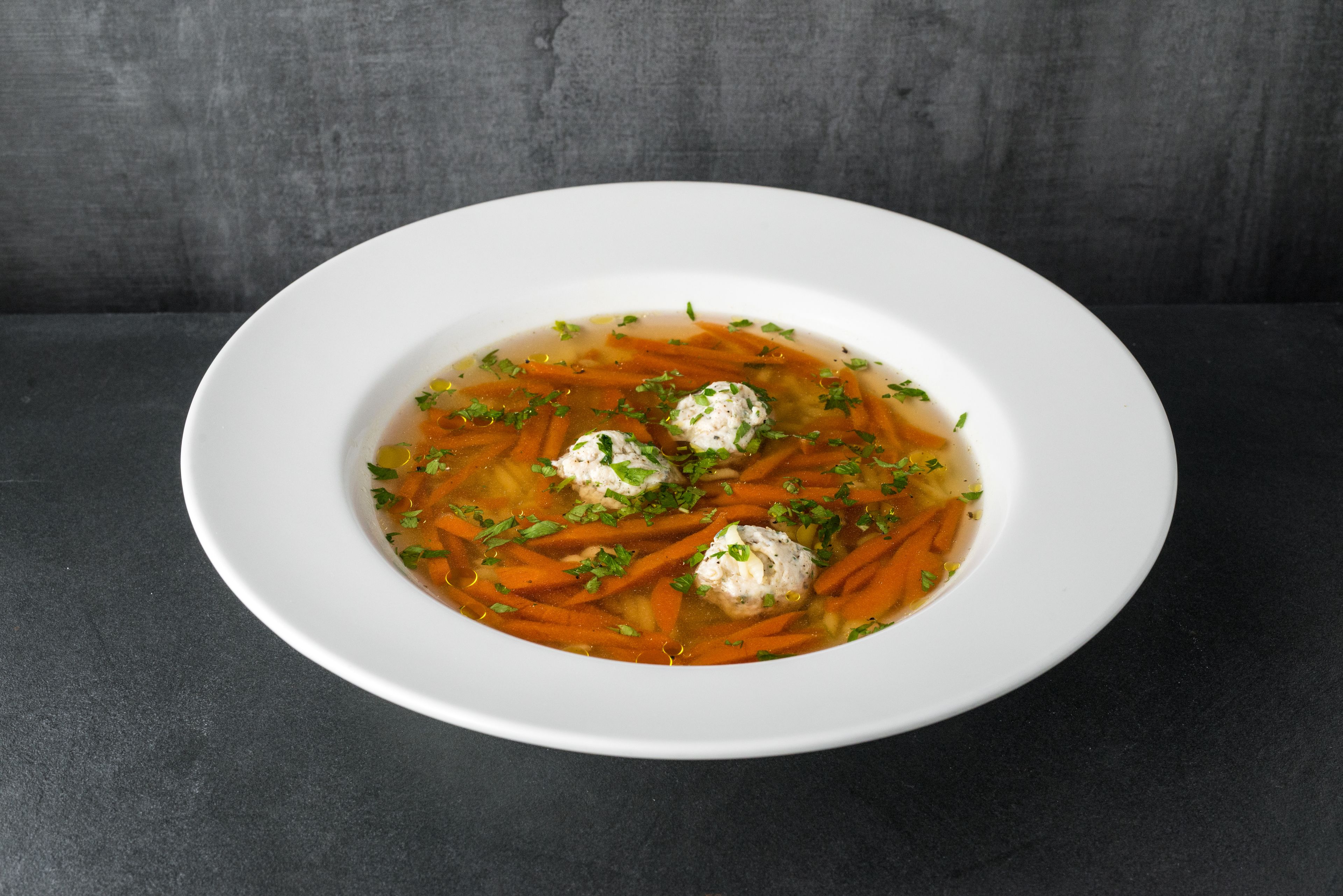 Une assiette ovale blanche contenant une soupe claire avec des carottes et des boulettes, décorée d’herbes, posée sur une surface sombre.