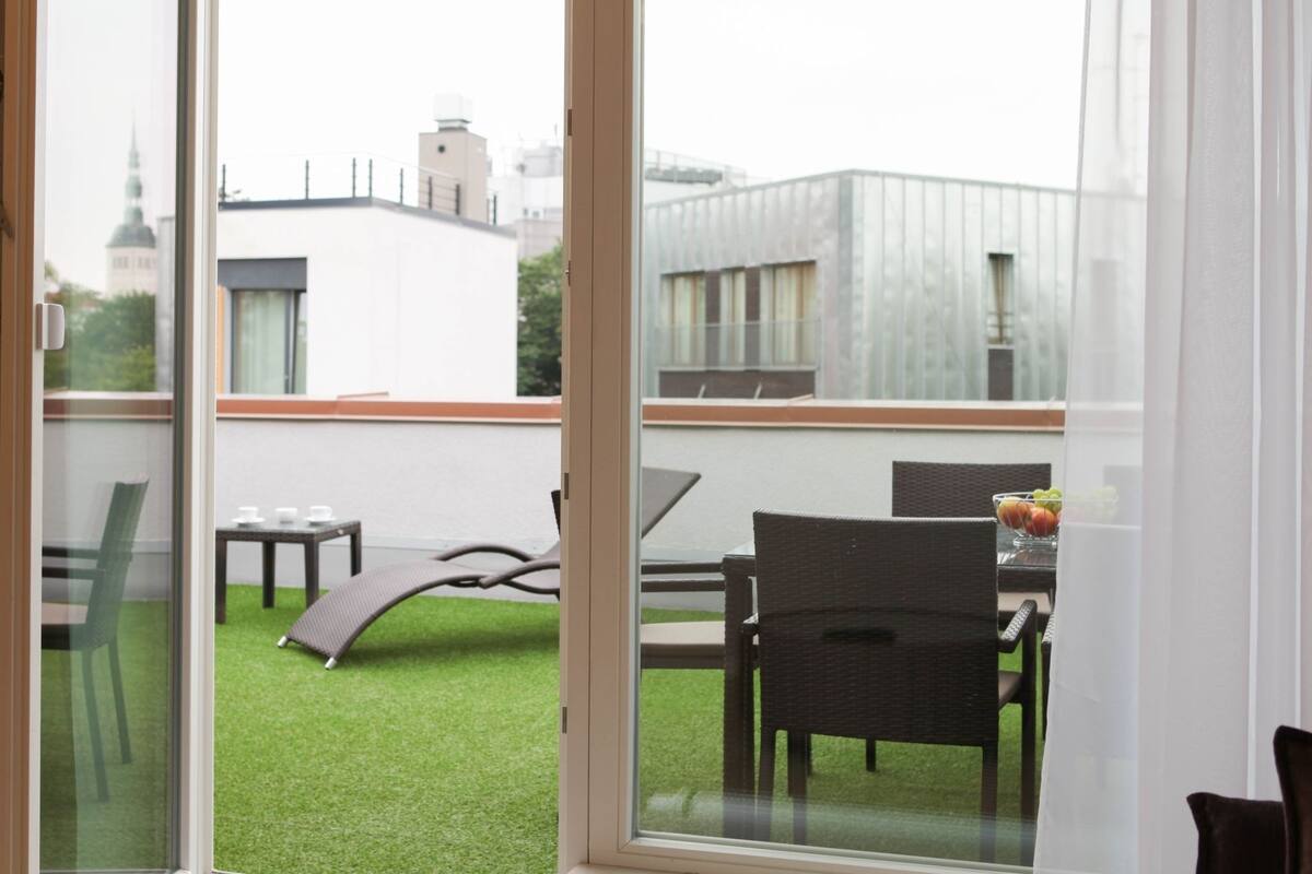 A balcony with a view of a cityscape, including a church spire. There are chairs, a table with a bowl of fruit, and a small table with cups.