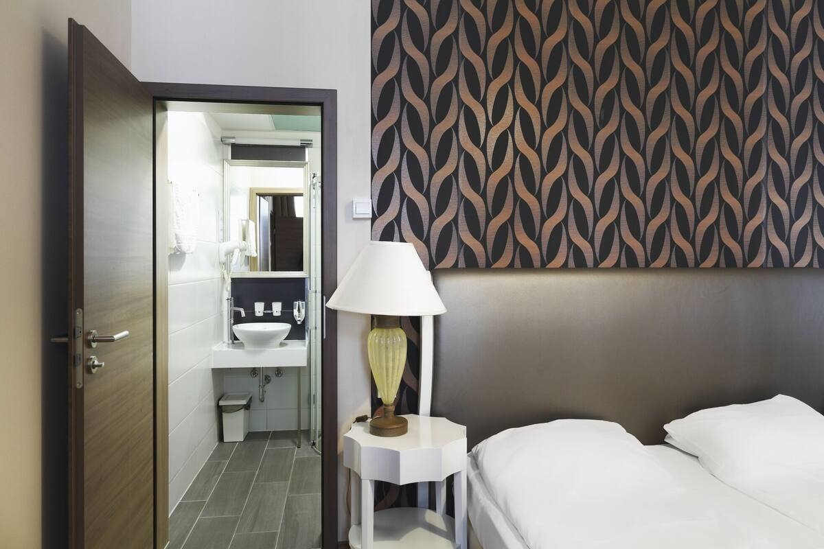 Ein Hotelzimmer mit Bett, Nachttisch mit Lampe, Badezimmer mit Waschbecken und Spiegel sowie einer Wand mit Muster.